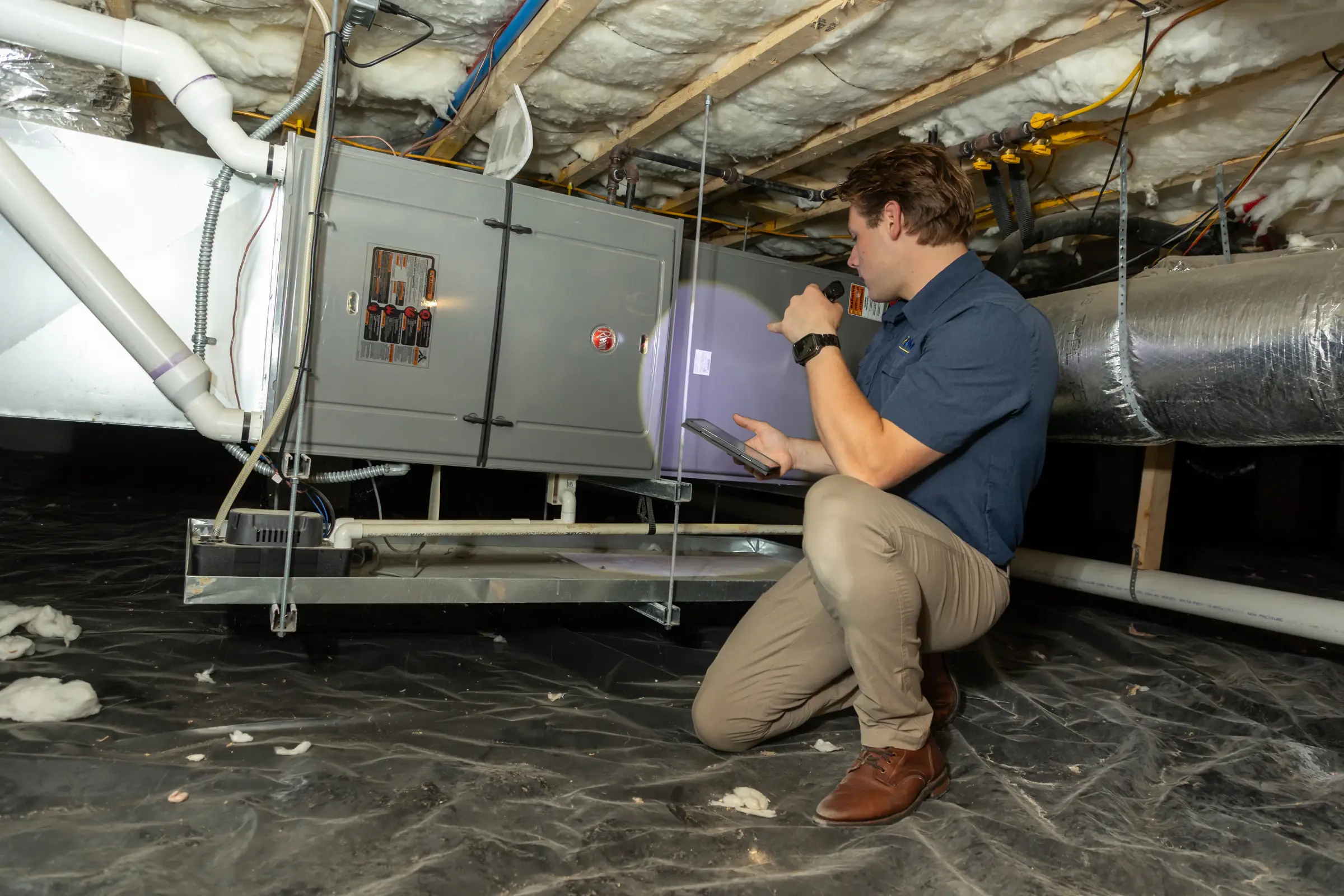 PMG technician checking an HVAC unit in a crawlspace with a flashlight and tablet